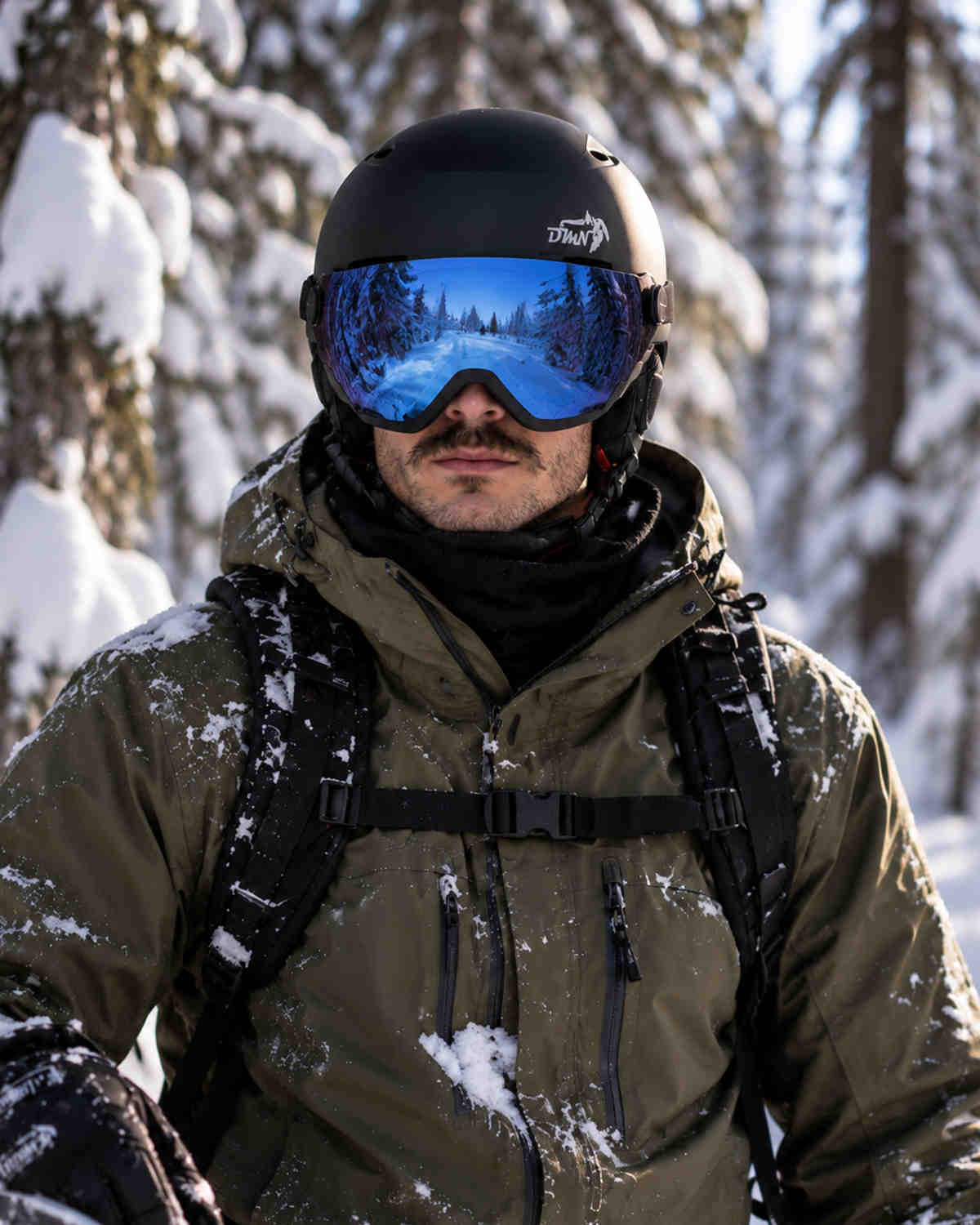 Vista frontale di snowboarder uomo con casco sci e snowboard NIVAL V DMN nero opaco e visiera blu specchiata integrata, durante una sessione freeride in un bosco innevato.