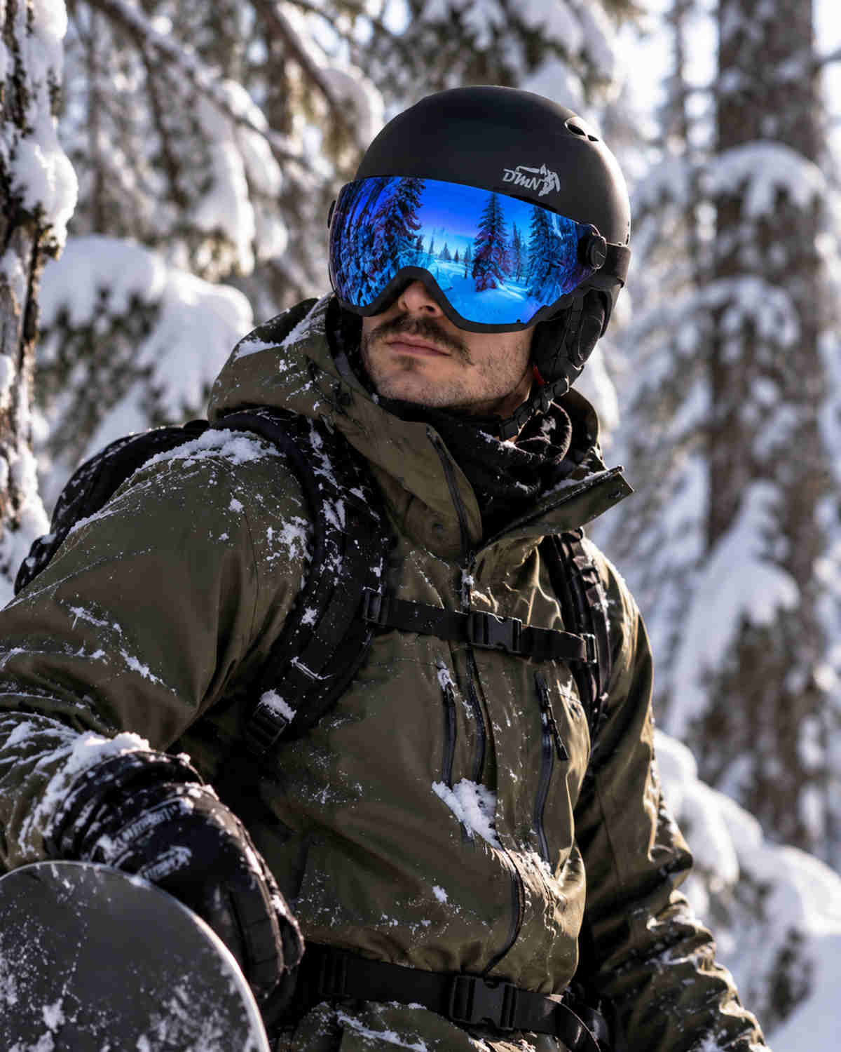 Snowboarder uomo con casco sci e snowboard NIVAL V DMN nero opaco con visiera blu specchiata integrata, durante una sessione freeride in un bosco innevato.