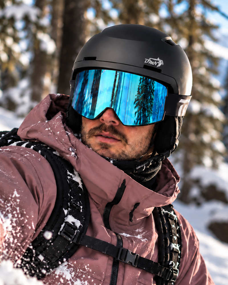 Uomo con casco snowboard DMN NIVAL nero opaco e maschera da neve blu specchiata, in un bosco innevato.